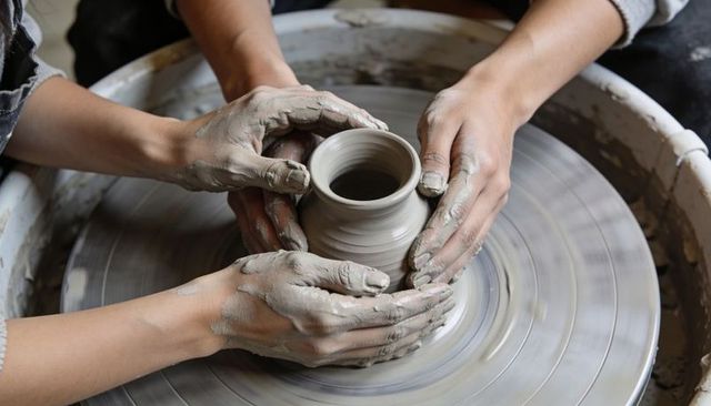 Mentor guiding student hands shaping small clay pot on pottery wheel in ceramics workshop