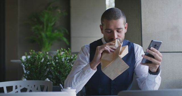 Male businessman is dressed in office attire, taking a break outdoors while eating from a paper bag. He is using a smartphone, showing the balance between work and rest. Useful for themes of workplace wellness, multitasking, and work-life balance.