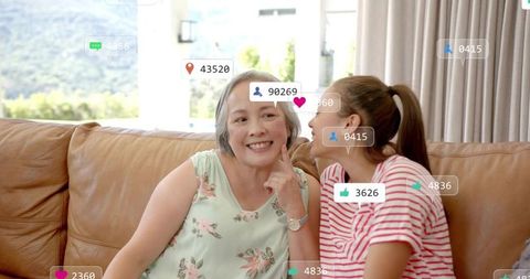 Smiling mother and daughter whispering on sunlit sofa with social media icons