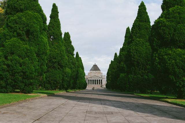 A serene pathway leads to a grand monument framed by majestic tall trees in a lush park. The overcast sky adds a calm, reflective mood to the scene. Ideal for use in travel blogs, brochures, and advertisements promoting outdoor recreation, tourism, or peaceful urban spaces.