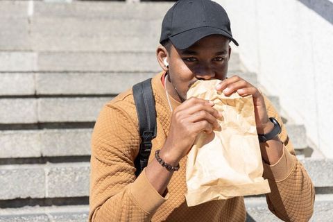 African american man sitting on city steps eating lunch from brown paper bag wearing cap