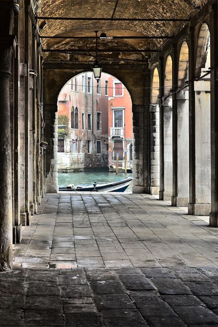 Archway leading to calm Venetian canal with a boat docked nearby. Arched structure with historic architecture detail. Ideal for travel blogs, architectural features studies, promoting Italian tourism or cultural explorations.