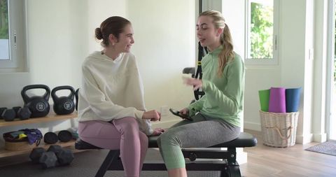 Female friends checking phone during home workout break on bench with kettlebells and mats