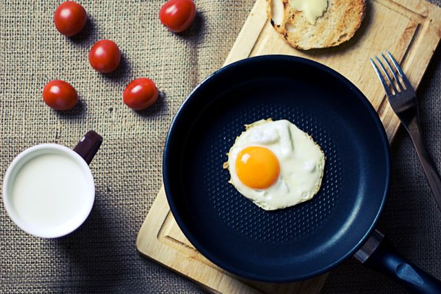 Sunny side up egg in a frying pan on wooden board with toast and cherry tomatoes. White cup with milk. Brown, textured background. Perfect for breakfast nutrition concept, cooking blogs, healthy lifestyle magazines.