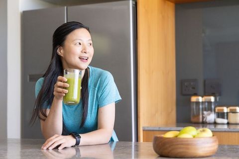 Mature asian woman enjoying green smoothie while leaning on modern kitchen counter