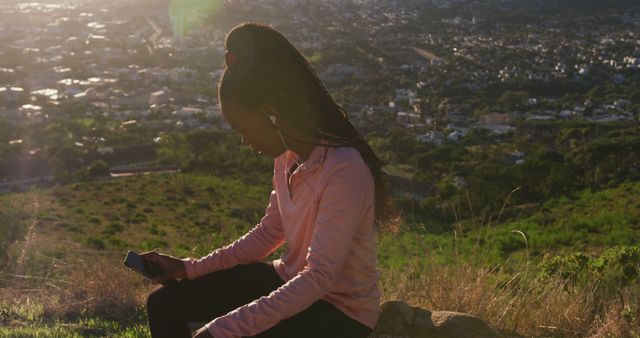 Young woman taking a break after exercise sitting on a hill with scenic cityscape in the background. Ideal for promoting fitness, outdoor activities, mindfulness, relaxation, and urban nature escapes.