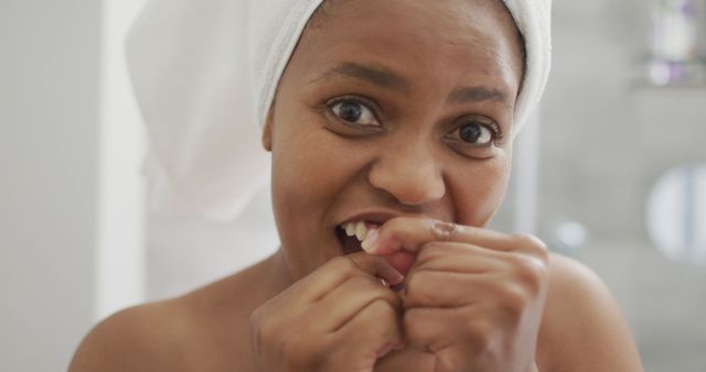 Young woman practicing oral hygiene by flossing teeth in the bathroom. She has a towel wrapped around her head, suggesting she just took a shower. Perfect for health, wellness, and self-care themes, as well as promoting dental products and routines.