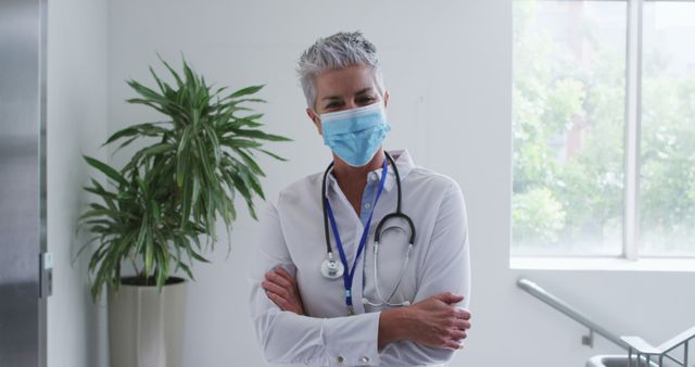 Portrait of caucasian female senior doctor wearing face mask standing with arms crossed in hospital. medicine, health and healthcare services during covid 19 coronavirus pandemic.