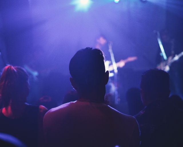 Crowd gathered, facing towards stage where performers are playing songs live. Blue lights illuminating stage create atmospheric mood. Perfect for promoting concerts, music events, nightlife activities, or entertainment marketing materials.