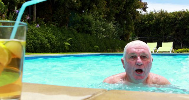 Senior man enjoying swimming in pool on a sunny day, with focus on man in tranquil garden environment. A cold drink with a straw is in the foreground, adding a refreshing touch to the scene. Perfect for concepts related to retirement, relaxation, summer vacations, outdoor enjoyment, and healthy lifestyle.