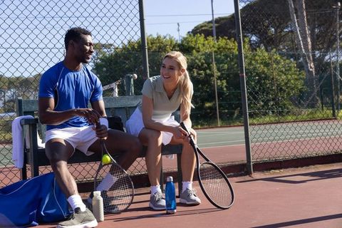 Tennis partners resting and chatting on court-side bench during sunny outdoor break