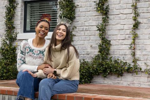 Smiling diverse women wearing merry headbands on cozy porch steps, festive winter friends