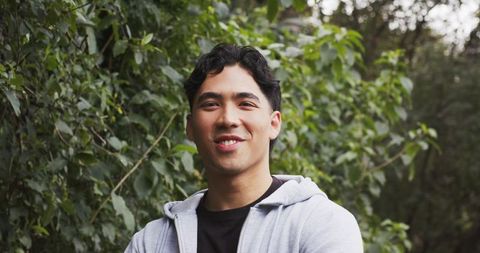 Young asian man standing and smiling in park wearing gray zip-up hoodie — outdoor portrait