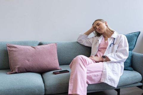 Tired female clinician resting on lounge sofa in pink scrubs with stethoscope and phone