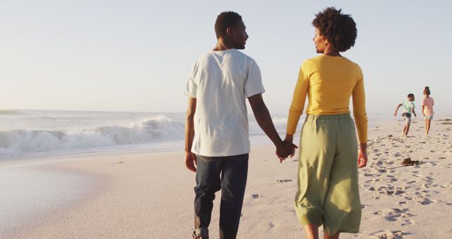 Smiling african american holding hands and walking on sunny beach. healthy, active family beach holiday.