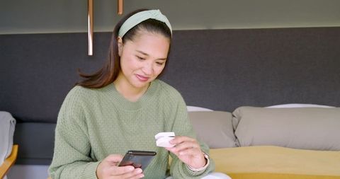 Asian woman in green sweater holding smartphone and earbud case