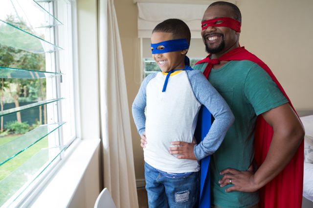 Father and son dressed in superhero costumes, standing by a window, smiling and looking outside. Ideal for themes of family bonding, playful moments, childhood imagination, and happy parenting. Perfect for use in advertisements, blogs, and articles about family life, parenting tips, and creative play.