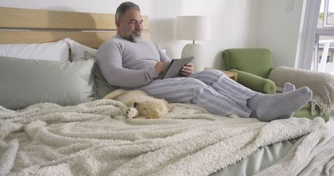 Middle-aged man relaxing with tablet and sleeping puppy in cozy bedroom