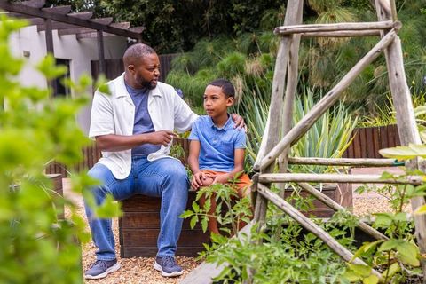 African american father talking with son sitting on wooden planter in backyard garden