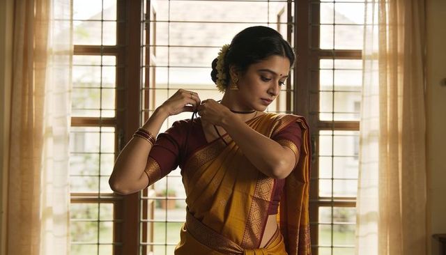 Indian woman fastening mangalsutra in mustard silk saree by wooden window, serene moment