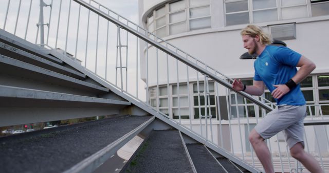 This image features a man running up a set of metal stairs outdoors near a modern building. He is wearing a blue t-shirt and grey shorts, emphasizing an active and healthy lifestyle. Perfect for use in marketing materials related to fitness, sports, urban living, and healthy lifestyle promotions.