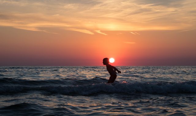 A child is playing in the ocean waves at sunset, captured as a silhouette against the vibrant sky. The sun is setting on the horizon, casting warm colors across the scene. Ideal for themes of childhood joy, beach vacations, summer activities, and serene natural beauty.