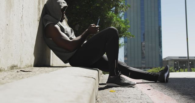 Side view of an African American man wearing a sleeveless hoodie and sportswear sitting on a step using a smartphone before training on a sunny day in the city.