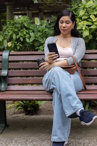 Young woman sitting on wooden park bench holding takeaway coffee and checking smartphone