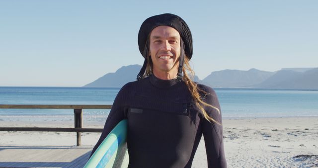 Image portrait of smiling caucasian man with dreadlocks in wetsuit holding surfboard on sunny beach. Freedom, sport, hobbies and healthy active lifestyle concept digitally generated image.