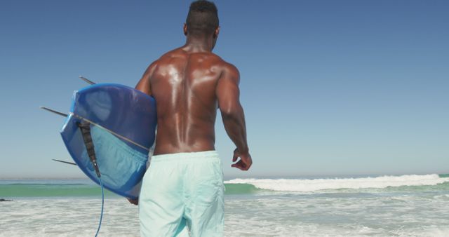 Man heading towards ocean holding a blue surfboard. Ideal for beach sports promotions, summer vacation ads, or surfing lifestyle features.