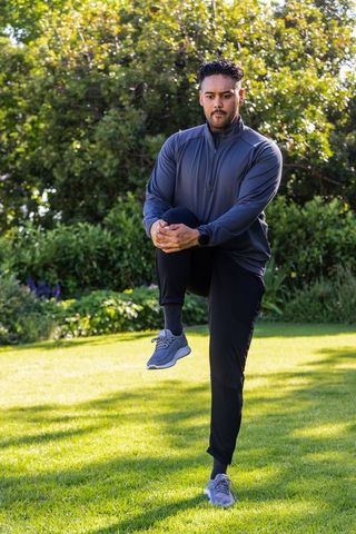 African american man performing knee hug stretch in sunlit garden morning workout with smartwatch