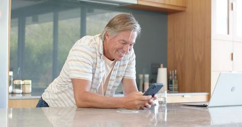 Mature man leaning on modern kitchen island checking smartphone while laptop sits open
