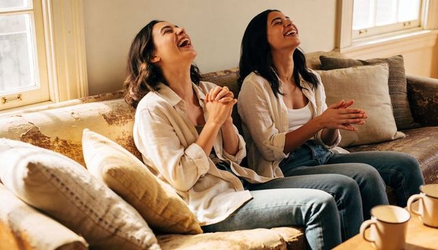 Laughing friends sharing coffee on sunlit cozy sofa, casual jeans and button-up shirts
