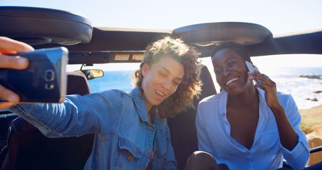 Two friends enjoying a scenic road trip by the coast. One is holding a smartphone to take a selfie while the other is on a phone call. Ideal for travel blogs, vacation advertisements, and social media campaigns promoting friendship and outdoor activities.