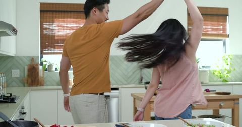 Diverse couple dancing in modern kitchen with wooden island, salad and smartphone