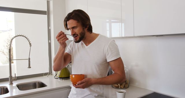 A man in a white t-shirt is eating cereal in a modern kitchen. He is holding an orange bowl and seems to be enjoying his breakfast. This image can be used for promoting healthy eating, breakfast ideas, kitchen appliances, or depicting daily morning routines in a household.