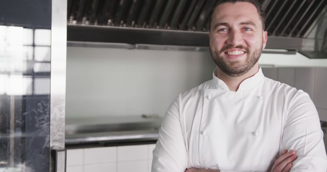 Smiling chef standing with arms crossed in a professional kitchen, wearing a white uniform. Useful for content related to culinary arts, restaurant business, hospitality industry, cooking skills, chef profiles, kitchen teamwork, and restaurant advertisements.