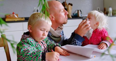 Father Caring for Daughter While Son Draws on Table at Home