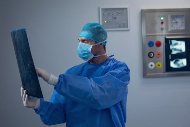 Male surgeon wearing blue surgical gown and mask carefully analyzing an x-ray in the hospital operating room. Relevant for medical, healthcare, and diagnostic applications. Could be used in medical websites, hospital brochures, and healthcare promotions.