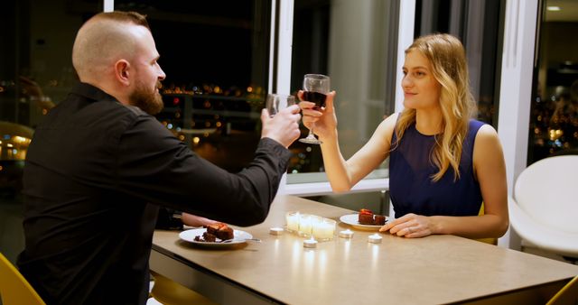 Couple enjoying a romantic dinner in a stylish, modern setting with candlelight. Both are toasting with glasses of wine and sharing dessert while surrounded by soft lighting and a city view through large windows. Suitable for themes related to romance, date nights, special occasions, anniversary celebrations, and intimate dining experiences.