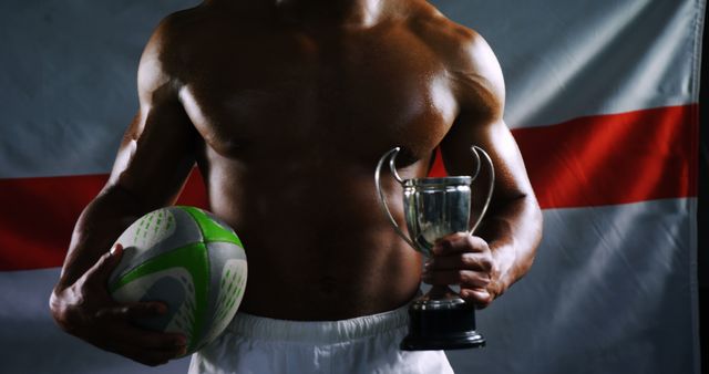 A shirtless, athletic young man holds a rugby ball and a trophy against a backdrop of the English flag, with copy space. His physique and the sports equipment suggest he is a rugby player celebrating a victory or achievement.