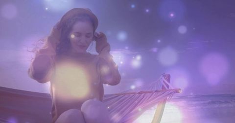 Adjusting beanie on hammock at twilight coastline with dreamy purple bokeh and serene mood