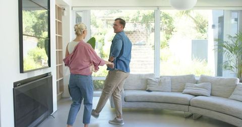 Young couple holding hands moving into modern sunlit living room with curved sofa