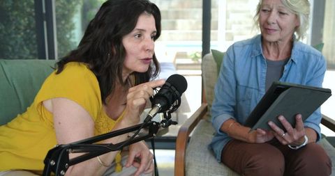 Mature women recording podcast together in home studio living room cohost interview using tablet