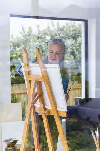 Senior woman painting at easel by window with natural light and garden reflections