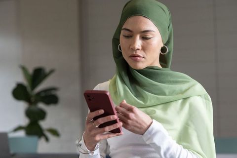 Woman wearing green hijab using smartphone in modern workspace focused expression