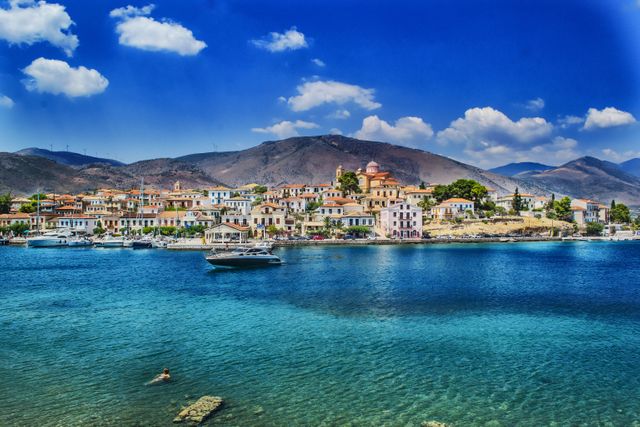 Depiction of a picturesque Greek coastal town with clear blue water and boats in the foreground. A swimmer is visible in the water, adding life to the serene scene. The town's buildings display traditional Mediterranean architecture, backdropped by hills under a vibrant blue sky with scattered clouds. This visual is perfect for tourism promotions, travel blogs, and vacation brochures, showcasing the beauty of Mediterranean destinations.