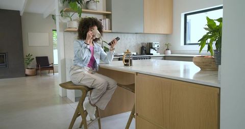 Woman sipping coffee and checking phone at modern sunlit kitchen island during morning