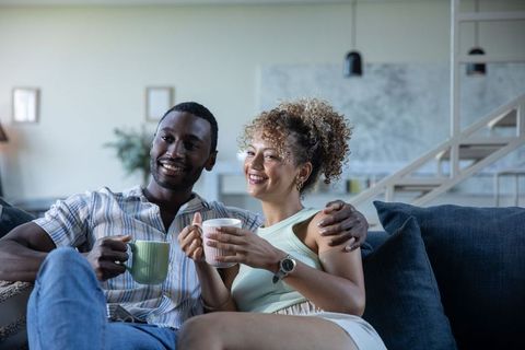 Multiracial couple relaxing on sofa holding mugs and smiling in cozy modern living room