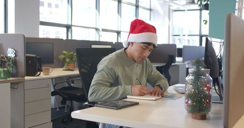 Young asian professional wearing santa hat writing notes at modern office with festive desk decor